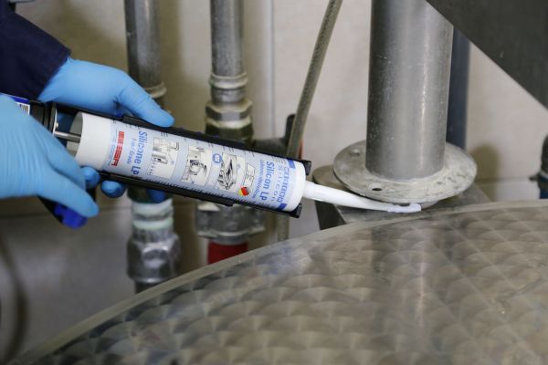 A person wearing blue gloves uses a caulking gun to apply WEICON Silicone LP around the flange joint of a metal pipe assembly.