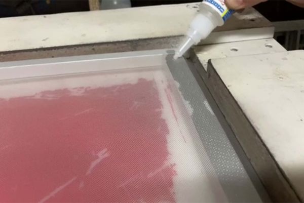 A person applies liquid adhesive from a small bottle along the edge of a screen printing frame.